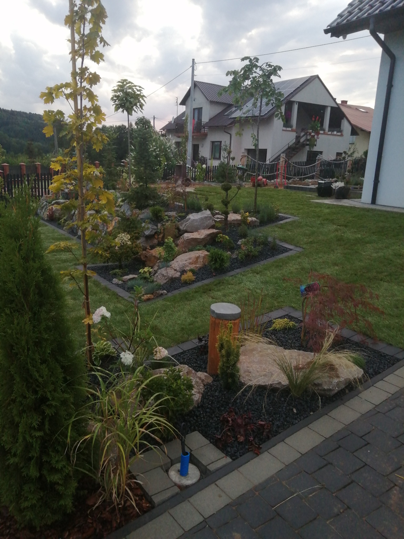 Uporządkowany ogród z równo przystrzyżonym trawnikiem, rabatami z kamieniami i roślinami ozdobnymi, widok z poziomu ścieżki z kostki brukowej.