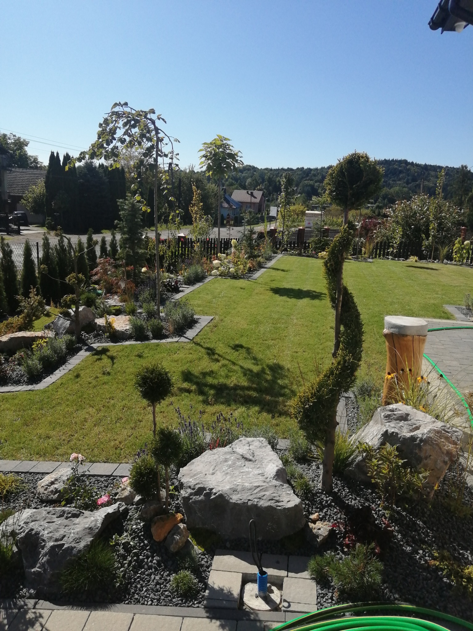 Ujęcie zadbanego ogrodu z trawnikiem, ozdobnymi drzewami formowanymi w kule i spiralę, rabatami z kamieniami i roślinami, oraz elementami małej architektury ogrodowej, jak pień z siedziskiem.