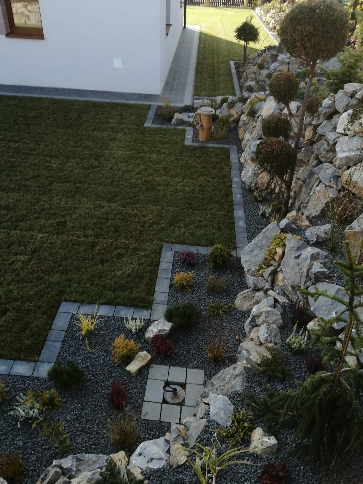 Ujęcie z góry na świeżo założony ogród z równo przystrzyżonym trawnikiem, skalniakiem z szarych kamieni i kulistymi drzewkami ozdobnymi, obrzeża z kostki brukowej oraz studzienka kanalizacyjna...