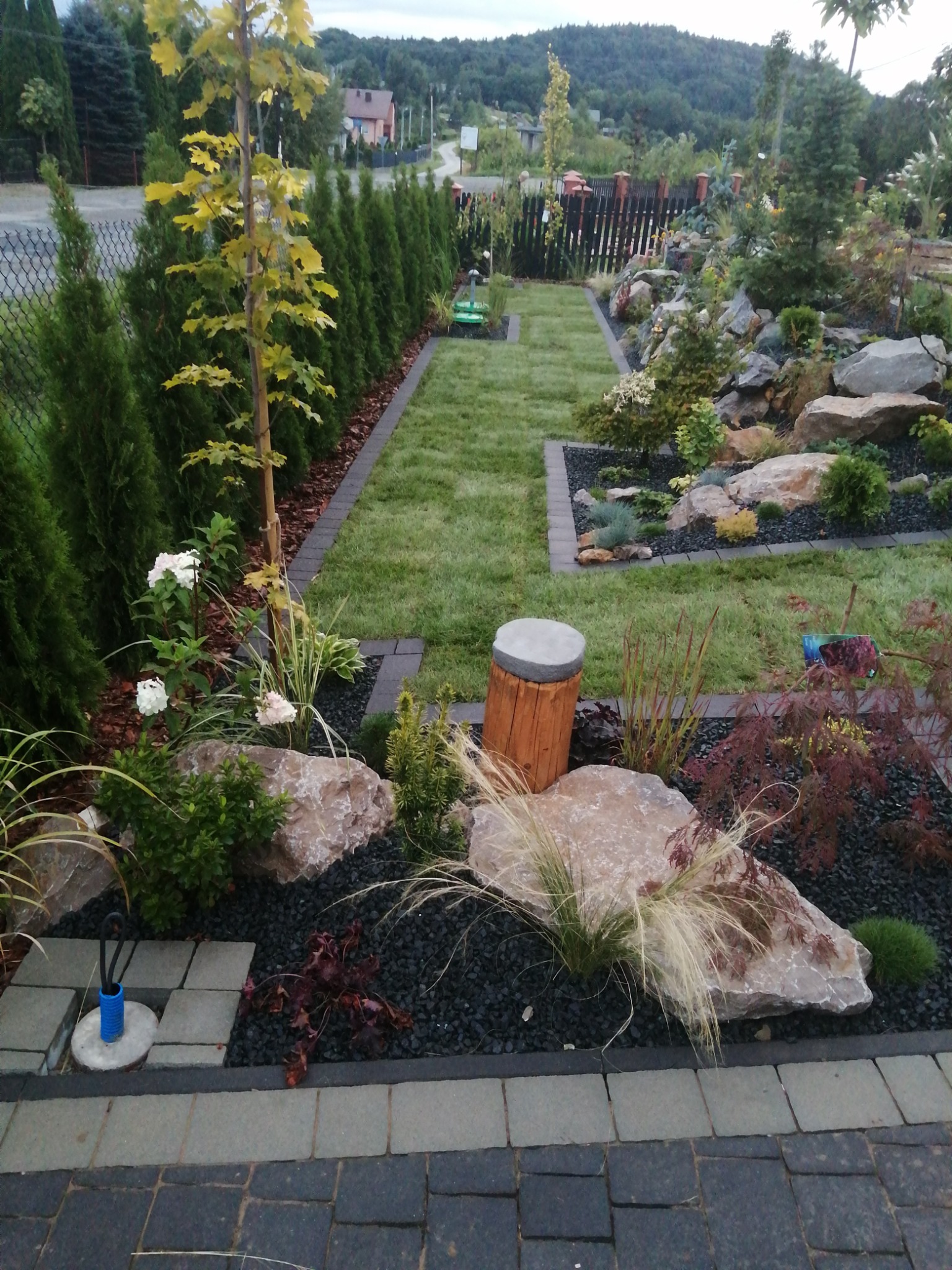 Ogród z trawnikiem, ścieżką z kostki, rabatami z kamieniami i roślinami ozdobnymi, w tle rząd tui i budynki mieszkalne.