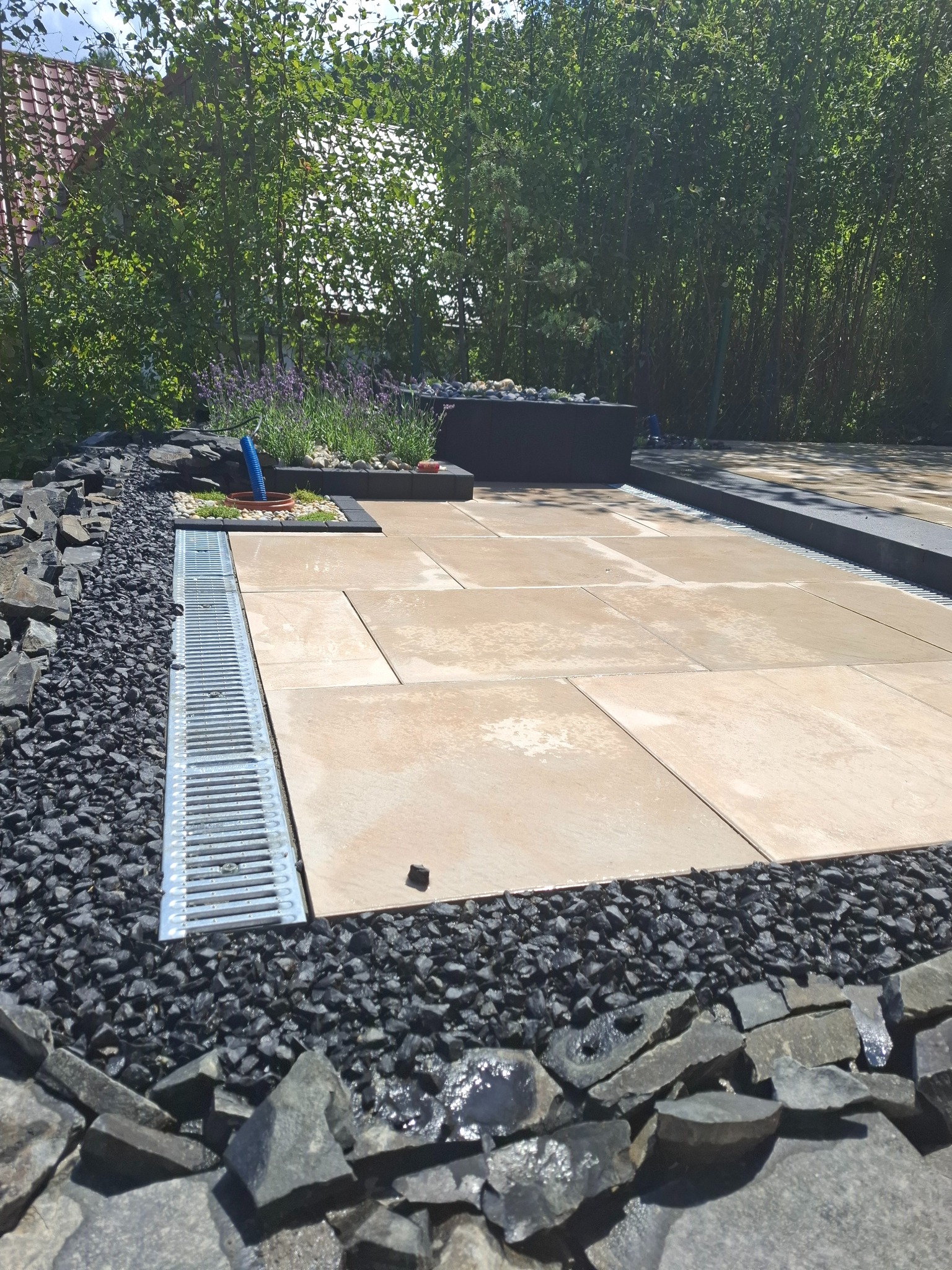 Nowoczesny taras z jasnych płyt, obramowany czarnym grysem i kamieniami, z systemem odprowadzania wody w postaci metalowej kratki. W tle widoczne drzewa i roślinność.