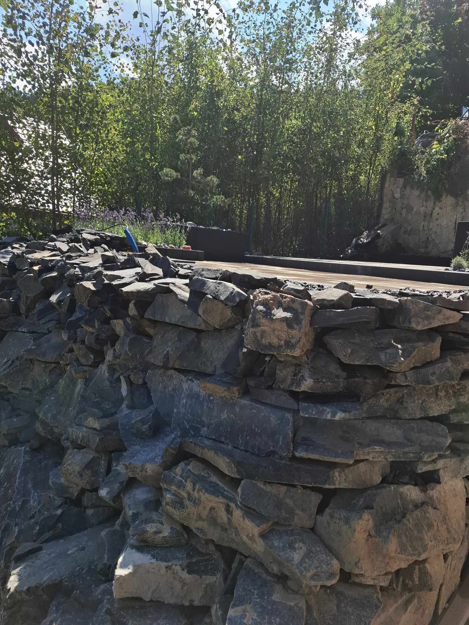 Kamienny murek z nieregularnych, szarych kamieni w ogrodzie, z widocznymi roślinami w tle i fragmentem drewnianego tarasu.