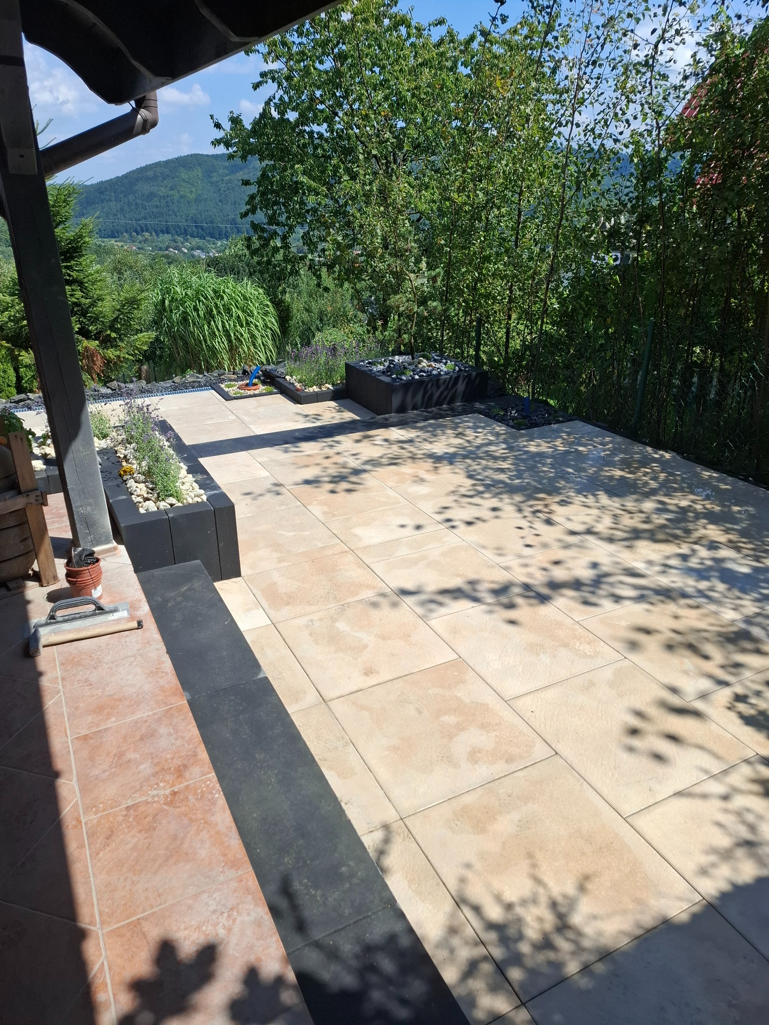 Wykończony taras z jasnych płyt z ciemnymi obrzeżami i podwyższonymi rabatami z lawendą i kamieniami, w tle zielony krajobraz z górami.