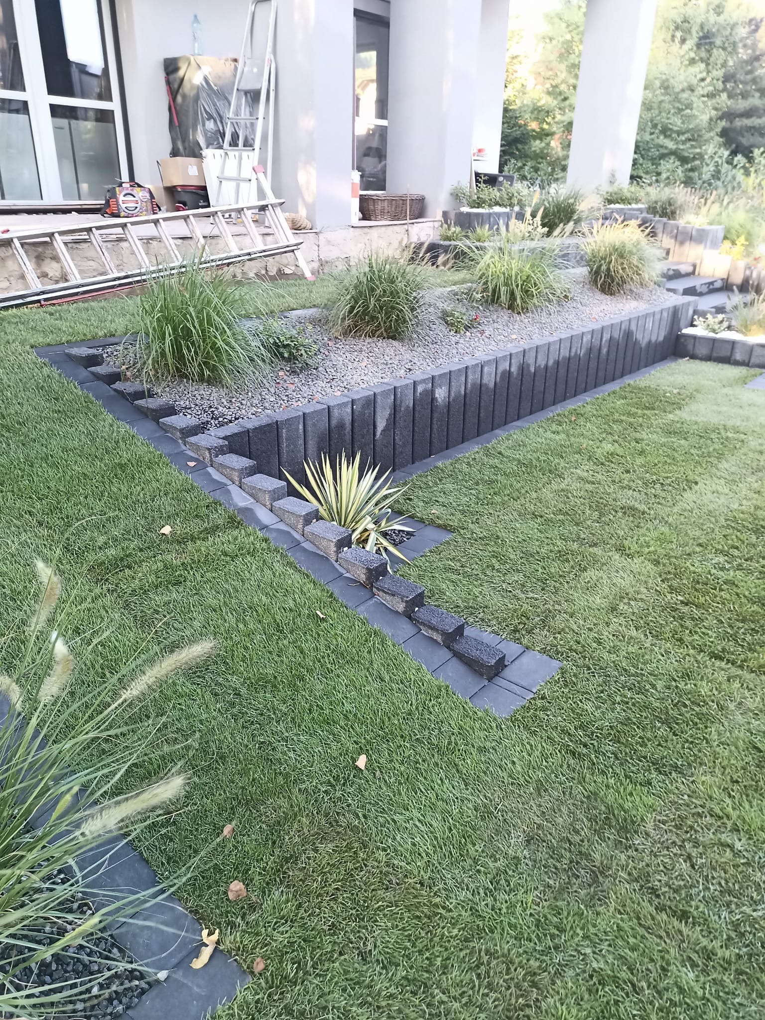 Nowoczesny ogród z geometrycznymi rabatami wykończonymi szarą kostką, ozdobne trawy i krzewy, trawnik, w tle biała elewacja domu i drabina.