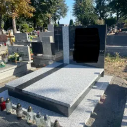 Nowoczesny, dwukolorowy pomnik z szarego i czarnego kamienia na cmentarzu, z zapalonymi zniczami u podstawy, w tle drzewa i inne nagrobki.
