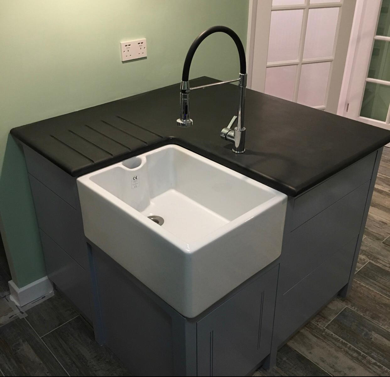 Biały, ceramiczny zlew farmerski wbudowany w ciemny blat z ociekaczem, bateria kuchenna z czarną wylewką, szare szafki kuchenne, podłoga imitująca drewno.