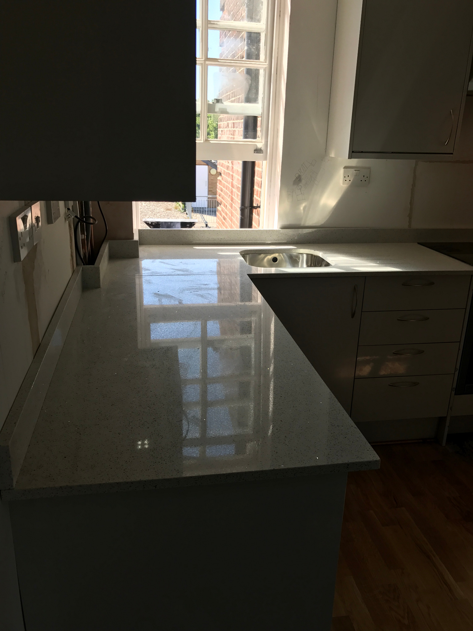 Jasny, błyszczący blat kuchenny z widocznym zlewem, odbijający światło z okna. Ściana z instalacją elektryczną i zarysami planowanych szafek. Na podłodze widoczny fragment drewnianego parkietu.