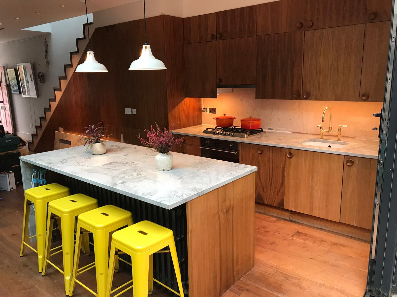 Nowoczesna kuchnia z drewnianymi szafkami i wyspą z blatem o marmurowym wzorze, uzupełniona czterema żółtymi stołkami barowymi i kwiatami w wazonie.