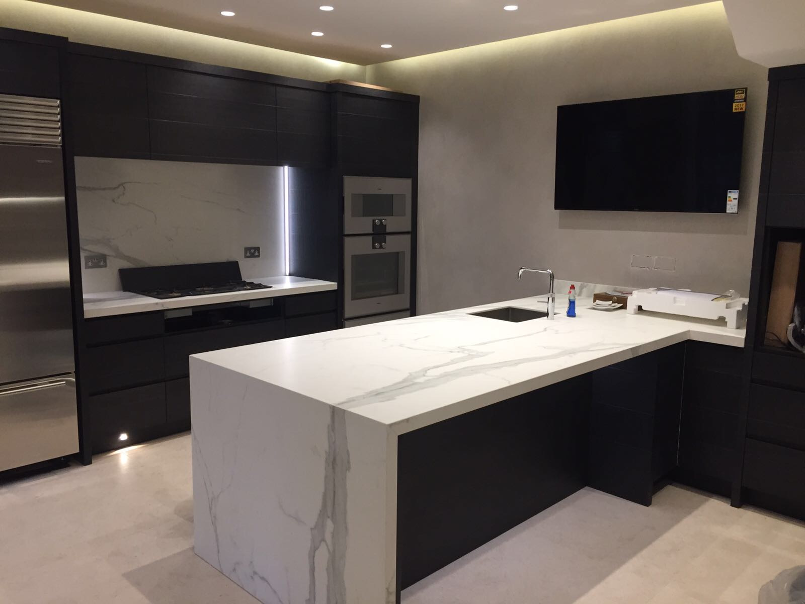 Nowoczesna kuchnia z czarnymi szafkami i blatem z białego kamienia z wyraźnym marmurkowym wzorem, oświetlenie LED podkreśla przestrzeń nad płytą grzewczą.