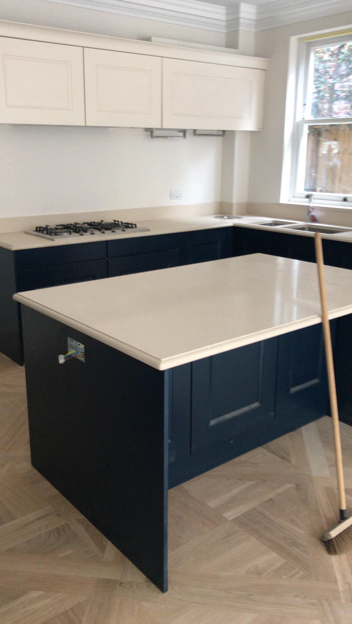 Nowoczesna kuchnia z ciemnoniebieską wyspą i blatem z jasnego kamienia, widoczne niedokończone podłączenie elektryczne, parkiet ułożony w jodełkę, w tle okno.