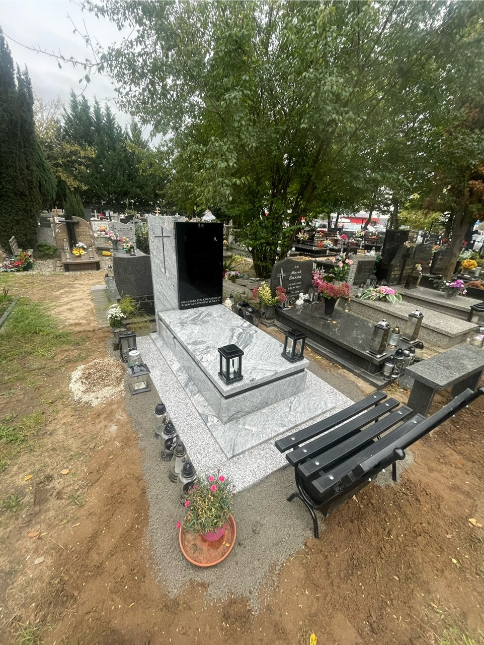 Jasnoszary pomnik z czarną tablicą, ozdobiony lampionami i kwiatami, ustawiony na cmentarzu obok czarnej ławki.