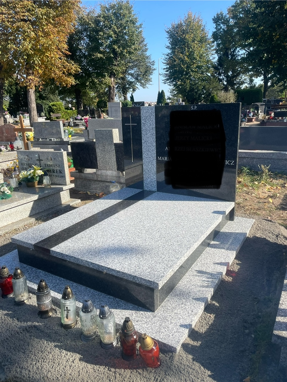 Nowoczesny, dwukolorowy pomnik z szarego i czarnego kamienia na cmentarzu, z zapalonymi zniczami u podstawy, w tle drzewa i inne nagrobki.