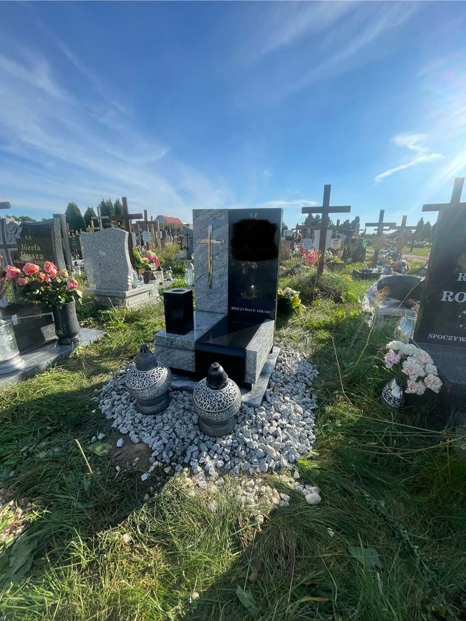 Czarny, granitowy pomnik z krzyżem na tle cmentarza z licznymi nagrobkami, otoczony jasnymi kamykami i dwoma ozdobnymi lampionami.