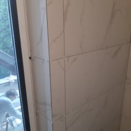 Katarzyna Wartecka - Świeżo ułożone, marmurowe płytki ścienne w rogu pomieszczenia, z widocznymi krzyżykami dystansowymi, z fragmentem okna i sylwetką pracownika w tle.