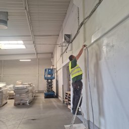 Katarzyna Wartecka - Dwóch pracowników w hali przemysłowej maluje ściany natryskowo, jeden z podnośnika, drugi z drabiny, na podłodze palety z materiałami budowlanymi.