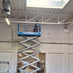 Katarzyna Wartecka - Pracownik na podnośniku maluje białą ścianę w hali magazynowej, widoczne instalacje pod sufitem i oświetlenie przemysłowe.