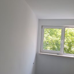 Katarzyna Wartecka - Świeżo pomalowane na biało wnętrze pokoju z oknem, przez które widać zielone drzewa, widoczny otwór w ścianie po lewej stronie okna.