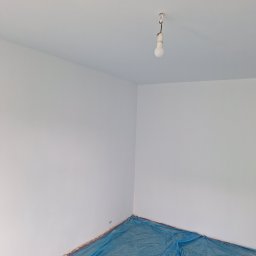 Katarzyna Wartecka - Pokój po świeżym malowaniu na biało, z widoczną żarówką na kablu zwisającą z sufitu i niebieską folią ochronną na podłodze przy ścianach.