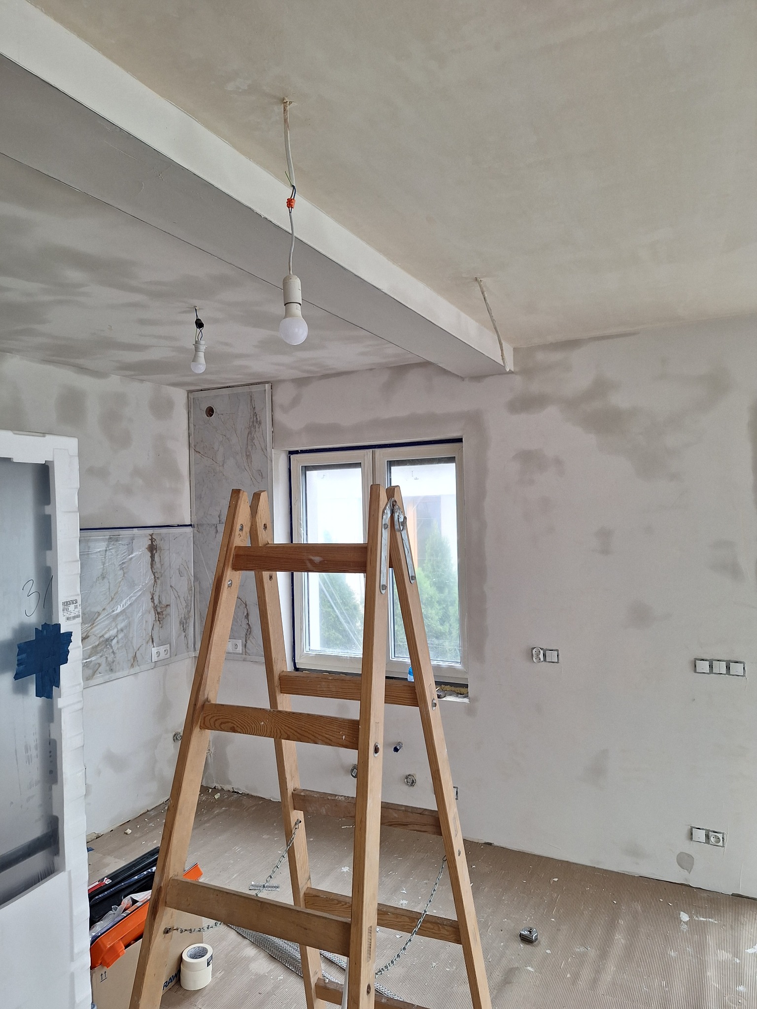 Pokój w trakcie szpachlowania ścian i sufitu, widoczna drabina drewniana, okno, instalacja elektryczna, fragment płytek imitujących marmur na ścianie oraz narzędzia malarskie na podłodze.