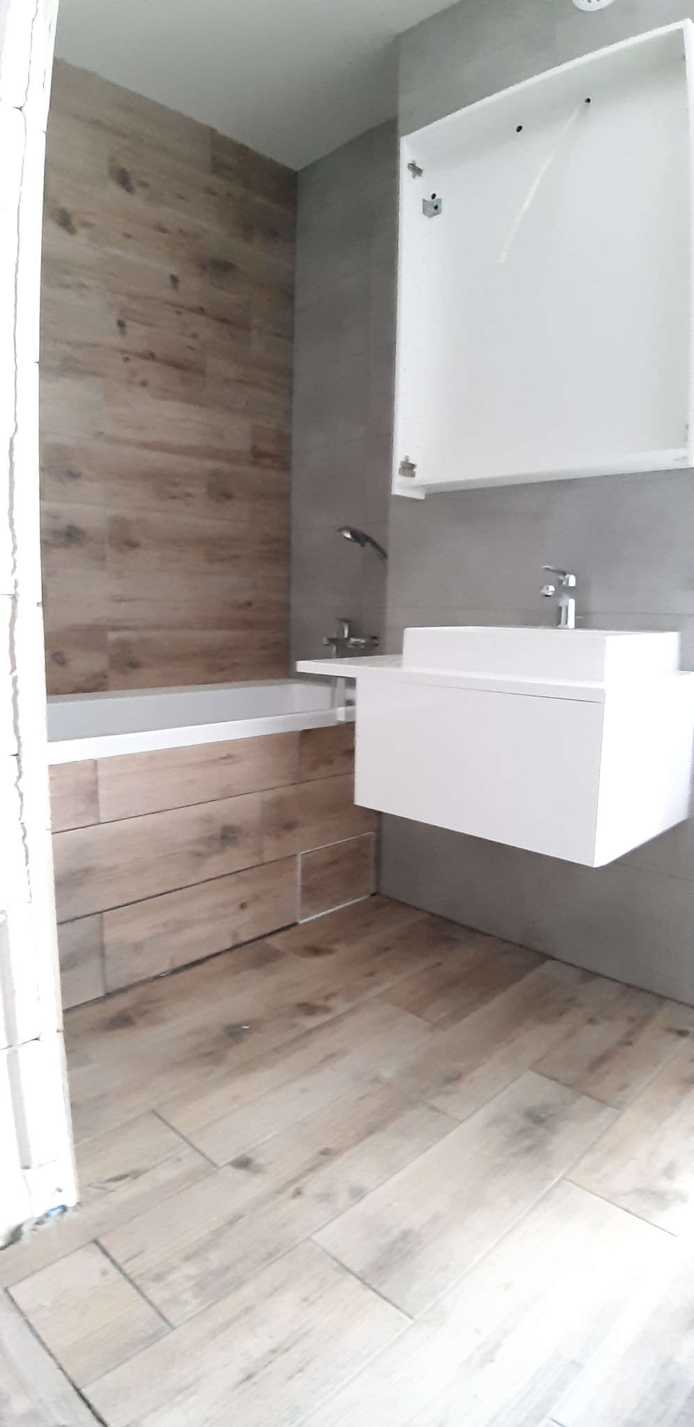 Nowoczesna łazienka z drewnopodobnymi płytkami na podłodze i ścianie wanny, białą szafką pod umywalką i wiszącą szafką z białymi frontami.