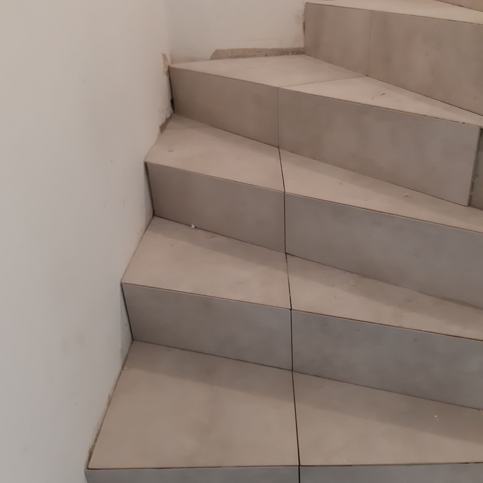 Kątowe ujęcie schodów wyłożonych płytkami w odcieniach beżu, widoczne niedoskonałości na styku ściany i pierwszego stopnia, sugerujące trwające prace wykończeniowe.