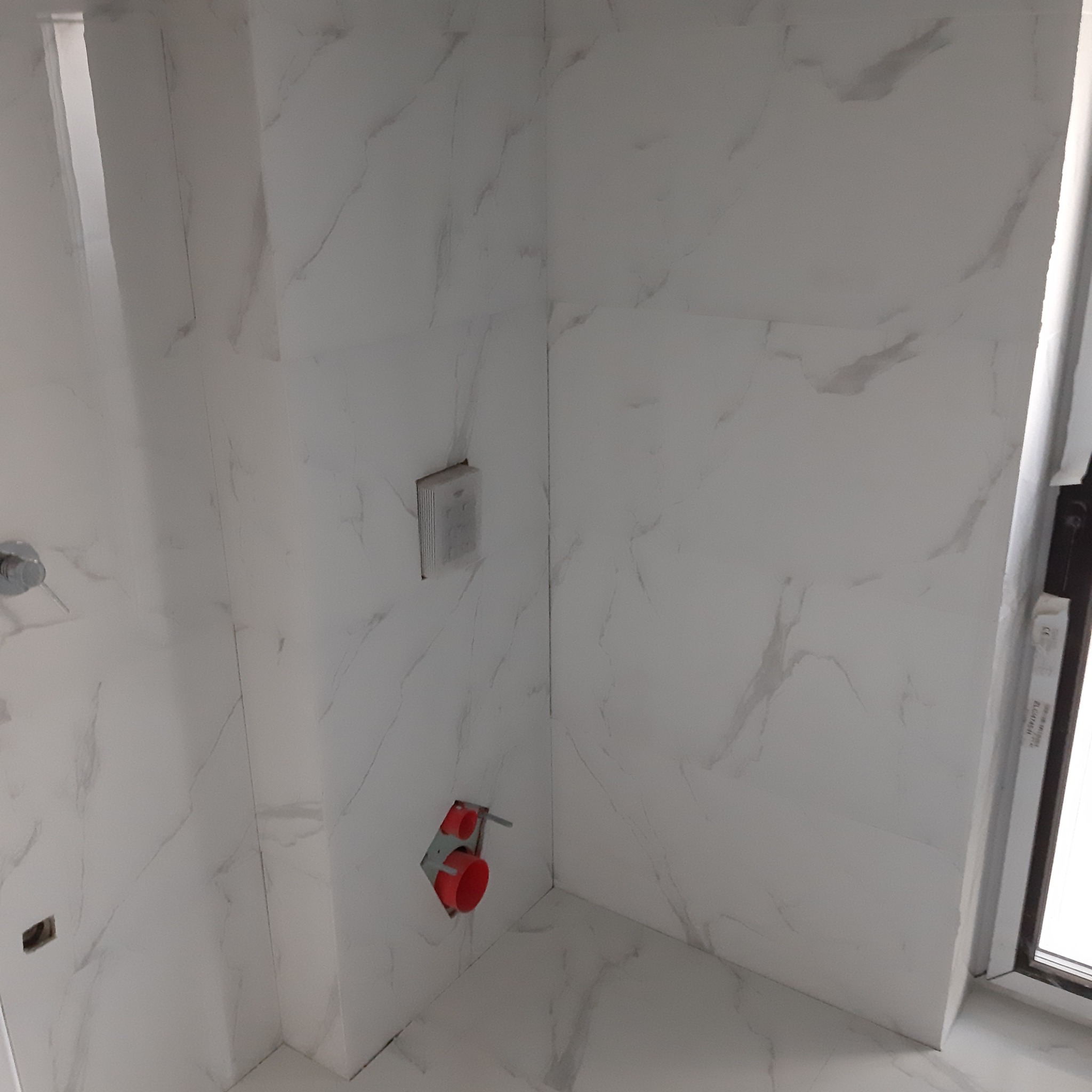 Ściana łazienki wyłożona białymi płytkami imitującymi marmur, z widocznymi elementami instalacji wodnej i elektrycznej przed montażem armatury.