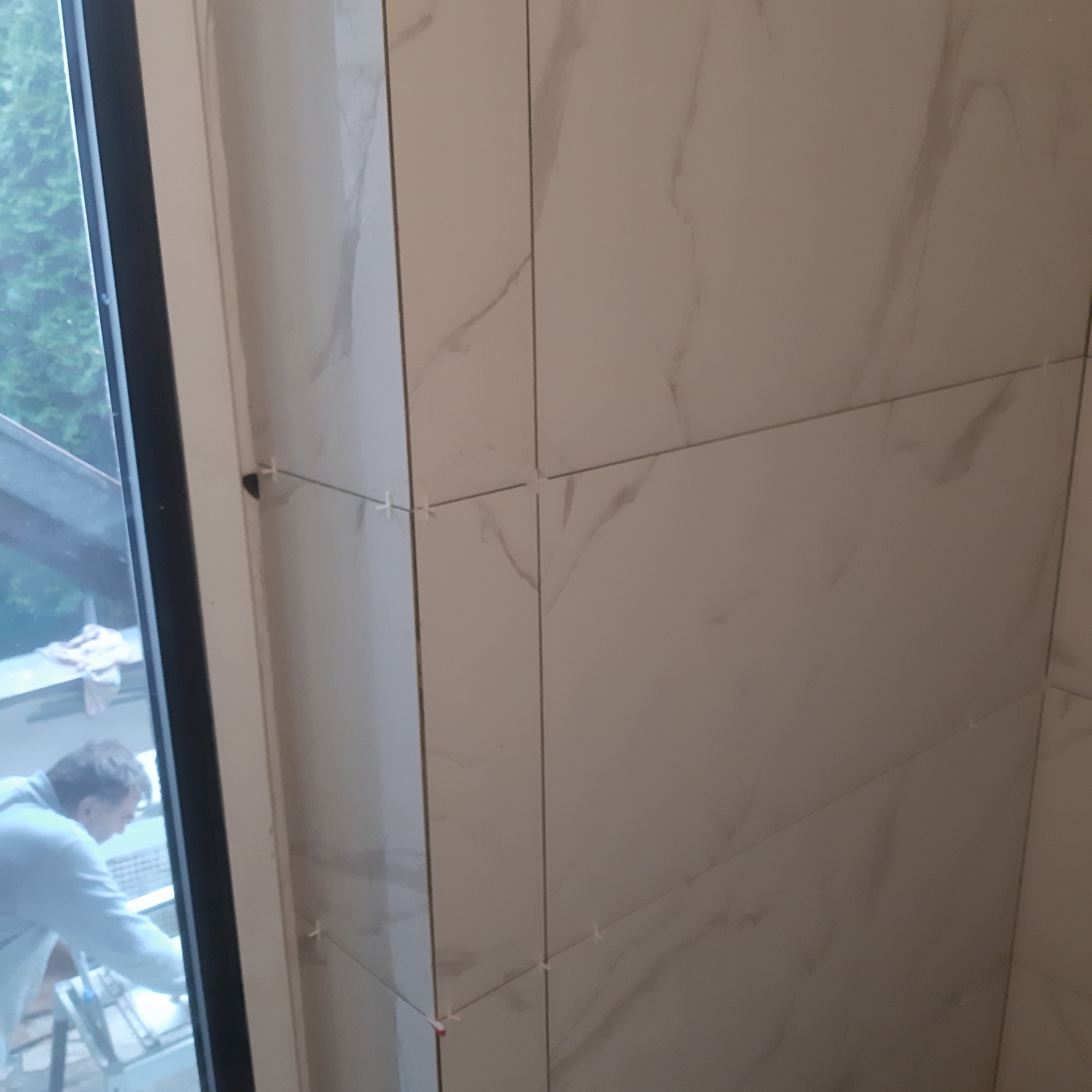 Świeżo ułożone, marmurowe płytki ścienne w rogu pomieszczenia, z widocznymi krzyżykami dystansowymi, z fragmentem okna i sylwetką pracownika w tle.