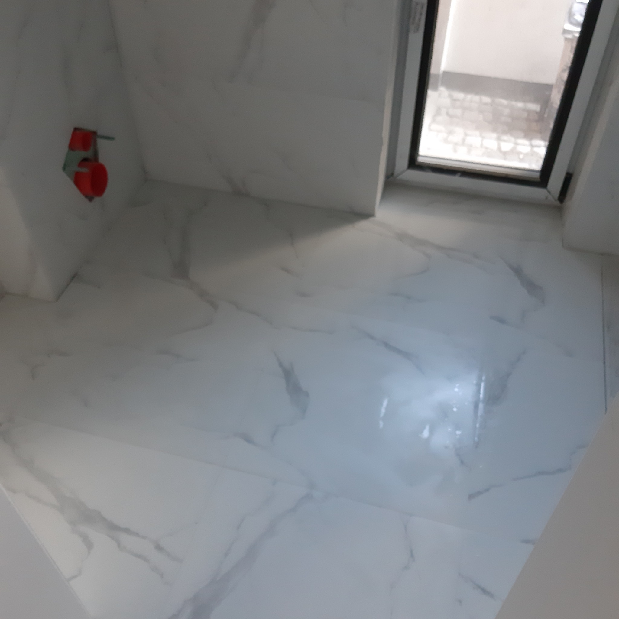 Wykończona łazienka z białymi płytkami imitującymi marmur na ścianach i podłodze, widoczne przyłącze wodne z czerwonymi elementami i okno z widokiem na zewnątrz.