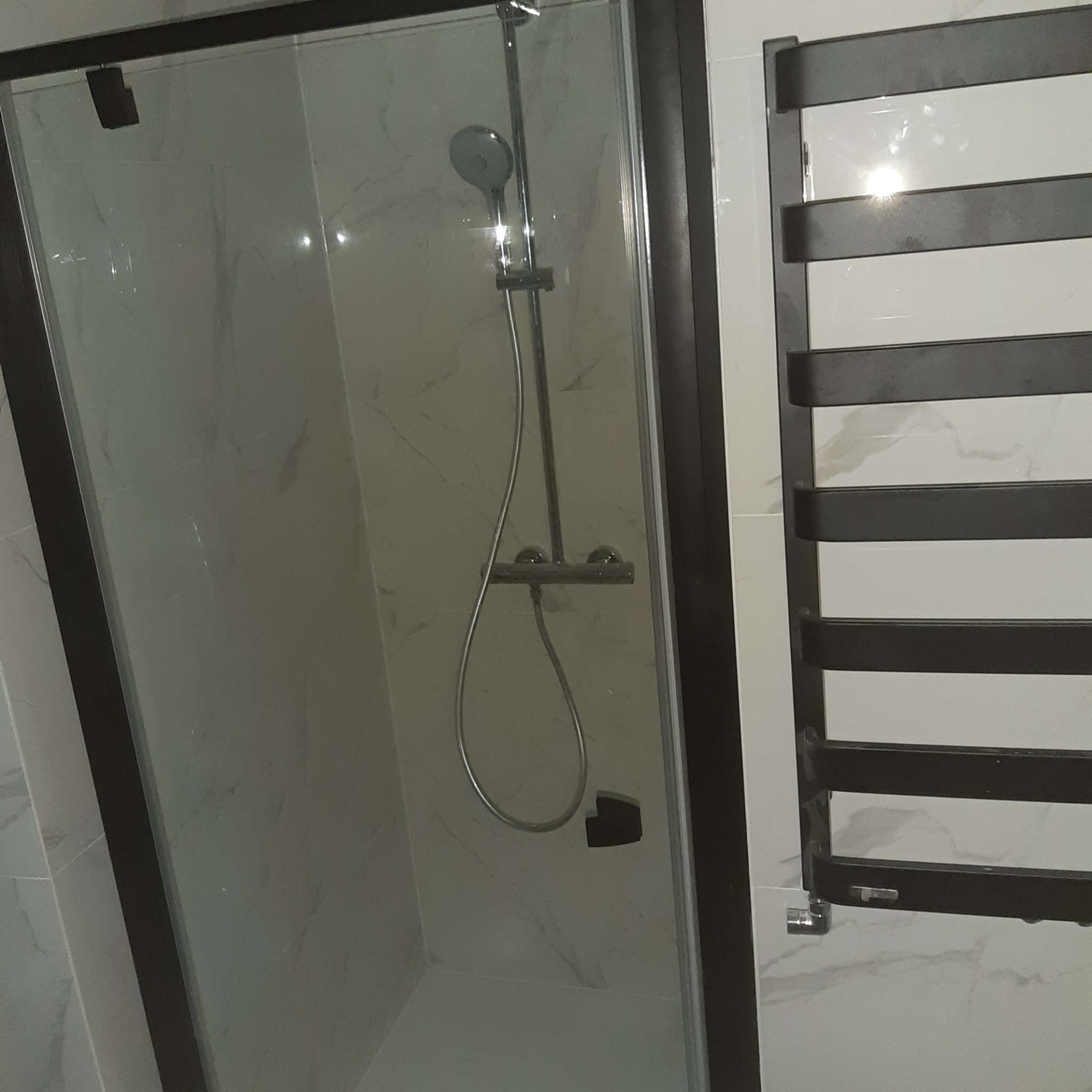 Nowoczesna kabina prysznicowa z czarną ramą i marmurowym wykończeniem ścian, widoczny grzejnik drabinkowy w kolorze antracytowym.