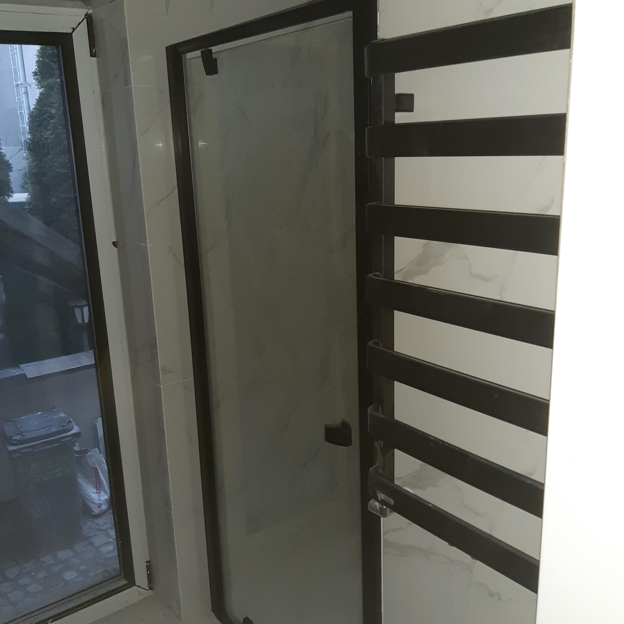 Zamknięta szklana kabina prysznicowa z ciemną ramą i grzejnikiem drabinkowym w łazience wykończonej płytkami imitującymi marmur.