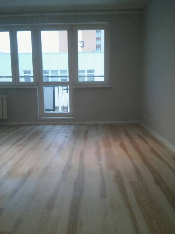 Jasne panele podłogowe ułożone w pustym pokoju z oknem balkonowym; widok na budynek z numerem '3'.