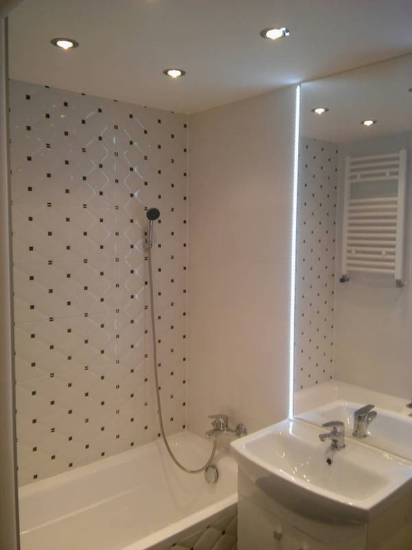 Biała łazienka z wanną i umywalką pod lustrem, ściany wyłożone płytkami w biało-czarną kratkę, oświetlenie punktowe w suficie.