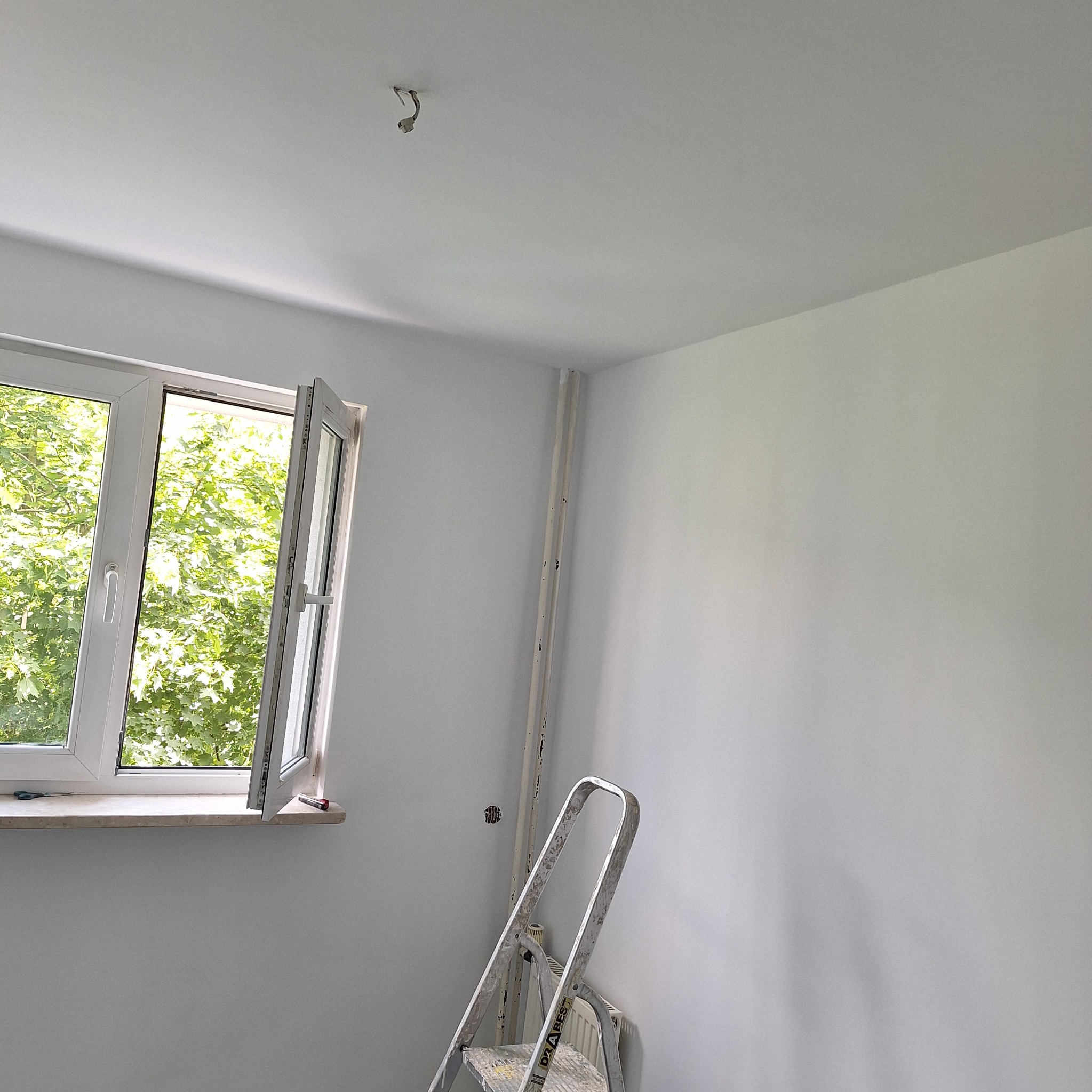 Świeżo pomalowane na biało wnętrze, widok na otwarte okno z zielenią na zewnątrz, drabina oparta o ścianę, widoczne ślady po gniazdku elektrycznym.
