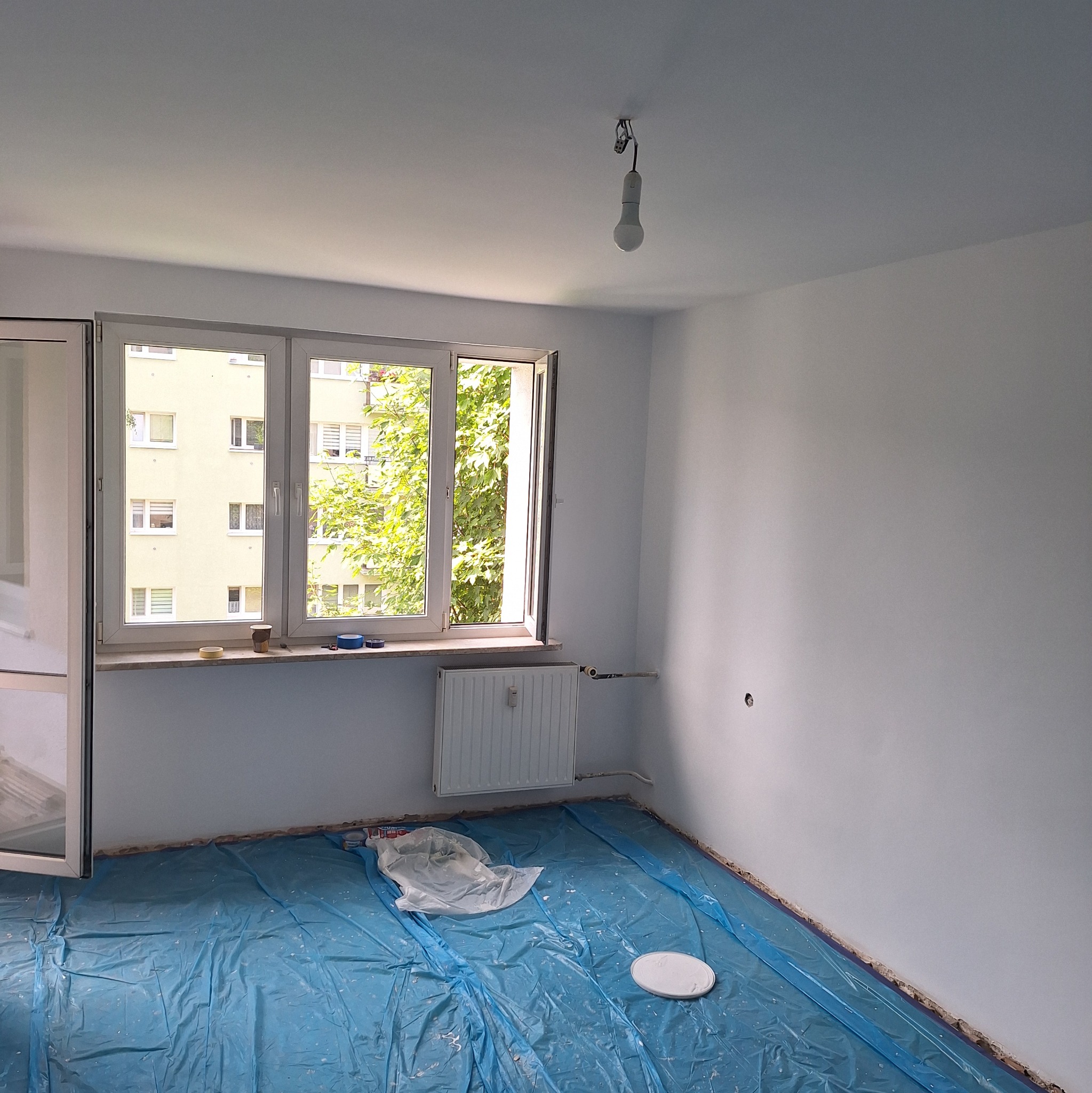 Pokój w trakcie odświeżania: ściany pomalowane na biało, podłoga zabezpieczona niebieską folią malarską, widoczne akcesoria malarskie i niedokończone prace przy listwach przypodłogowych.