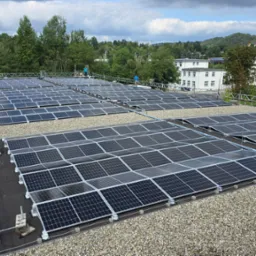 Widok z góry na rozległą instalację paneli słonecznych na dachu budynku, z pracownikami w niebieskich koszulkach na rusztowaniu, otoczoną zielenią drzew i zabudowaniami w tle.