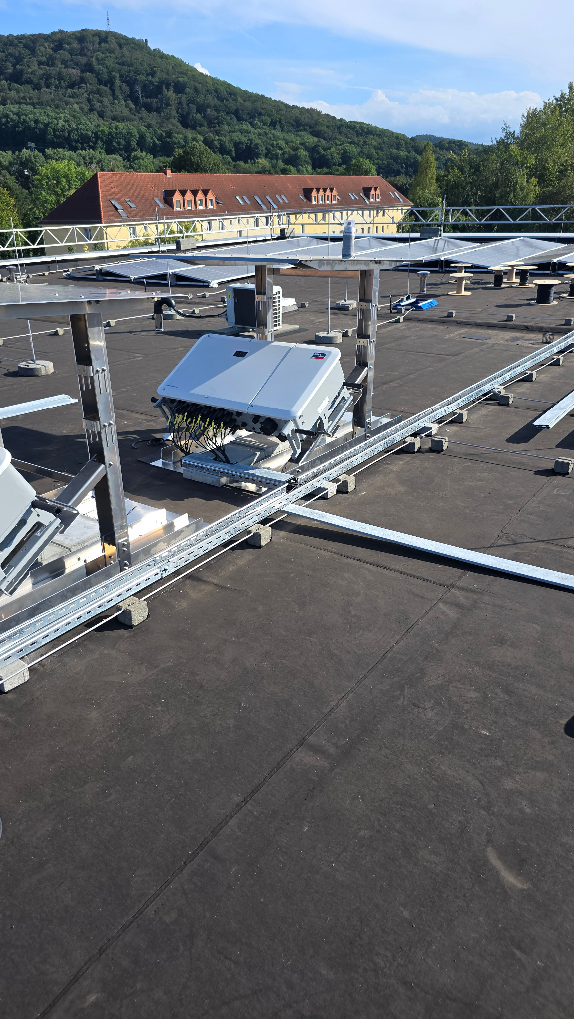 Szary inwerter solarny SMA zamontowany na dachu budynku z widocznymi kablami, szynami montażowymi i betonowymi podstawami, w tle budynek z czerwonym dachem i zalesione wzgórze.