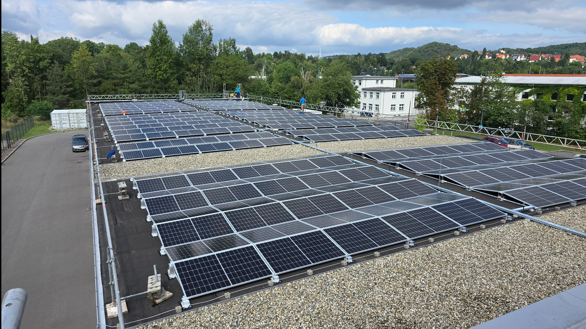 Widok z góry na rozległą instalację paneli słonecznych na dachu budynku, z pracownikami w niebieskich koszulkach na rusztowaniu, otoczoną zielenią drzew i zabudowaniami w tle.