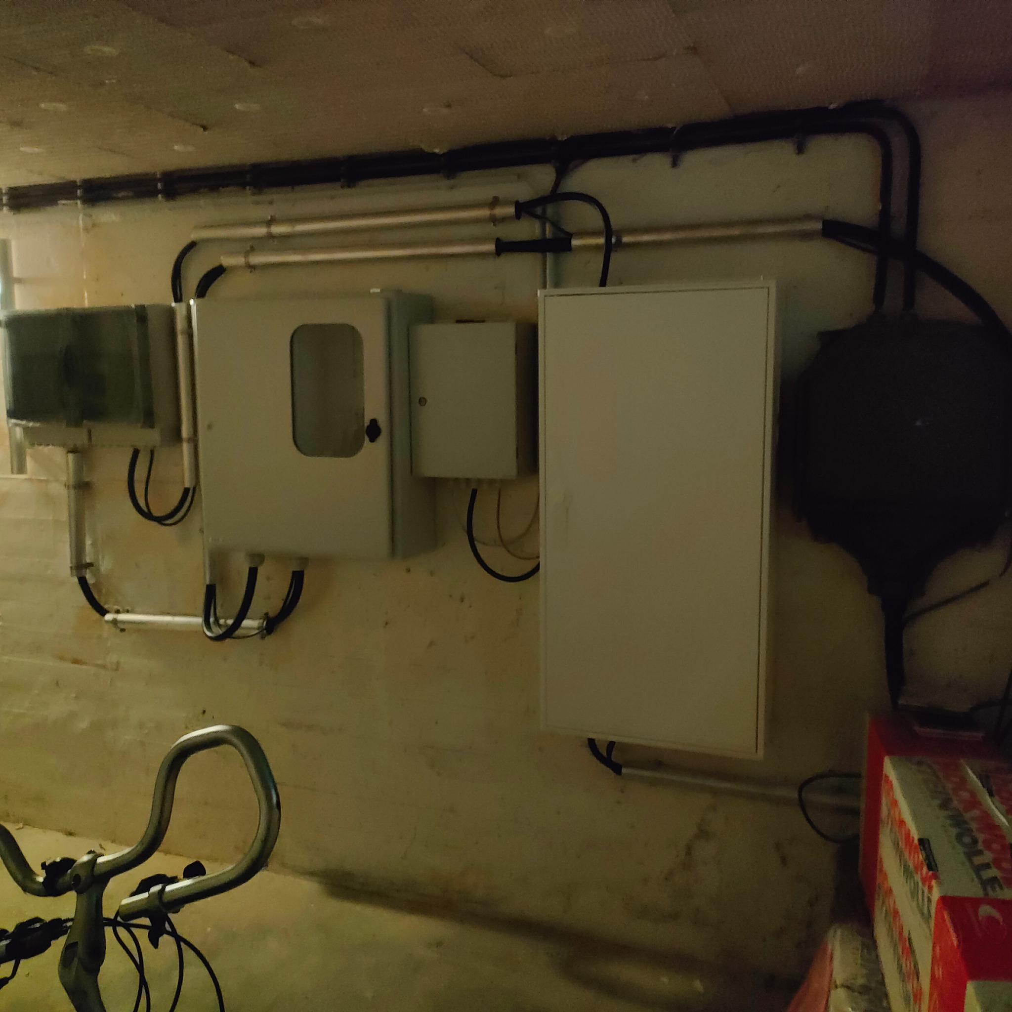 Szara ściana piwnicy z zamontowanymi skrzynkami elektrycznymi, rurami i przewodami; w tle widoczne opakowania izolacji ROCKWOOL oraz fragment kierownicy roweru.