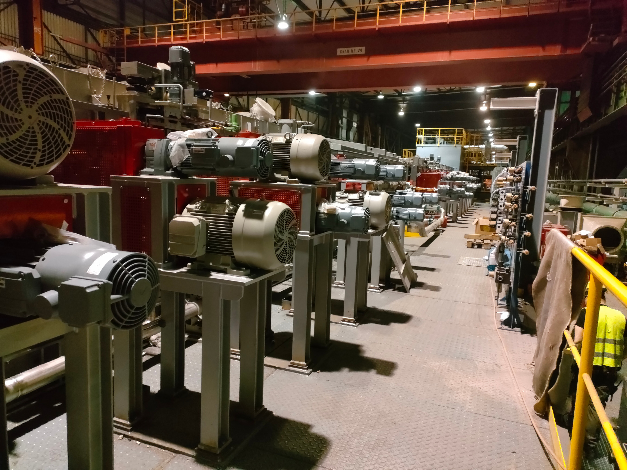 Rząd identycznych silników elektrycznych na metalowych podstawach w hali przemysłowej, widoczne czerwone skrzynki i żółta barierka, oświetlenie górne.