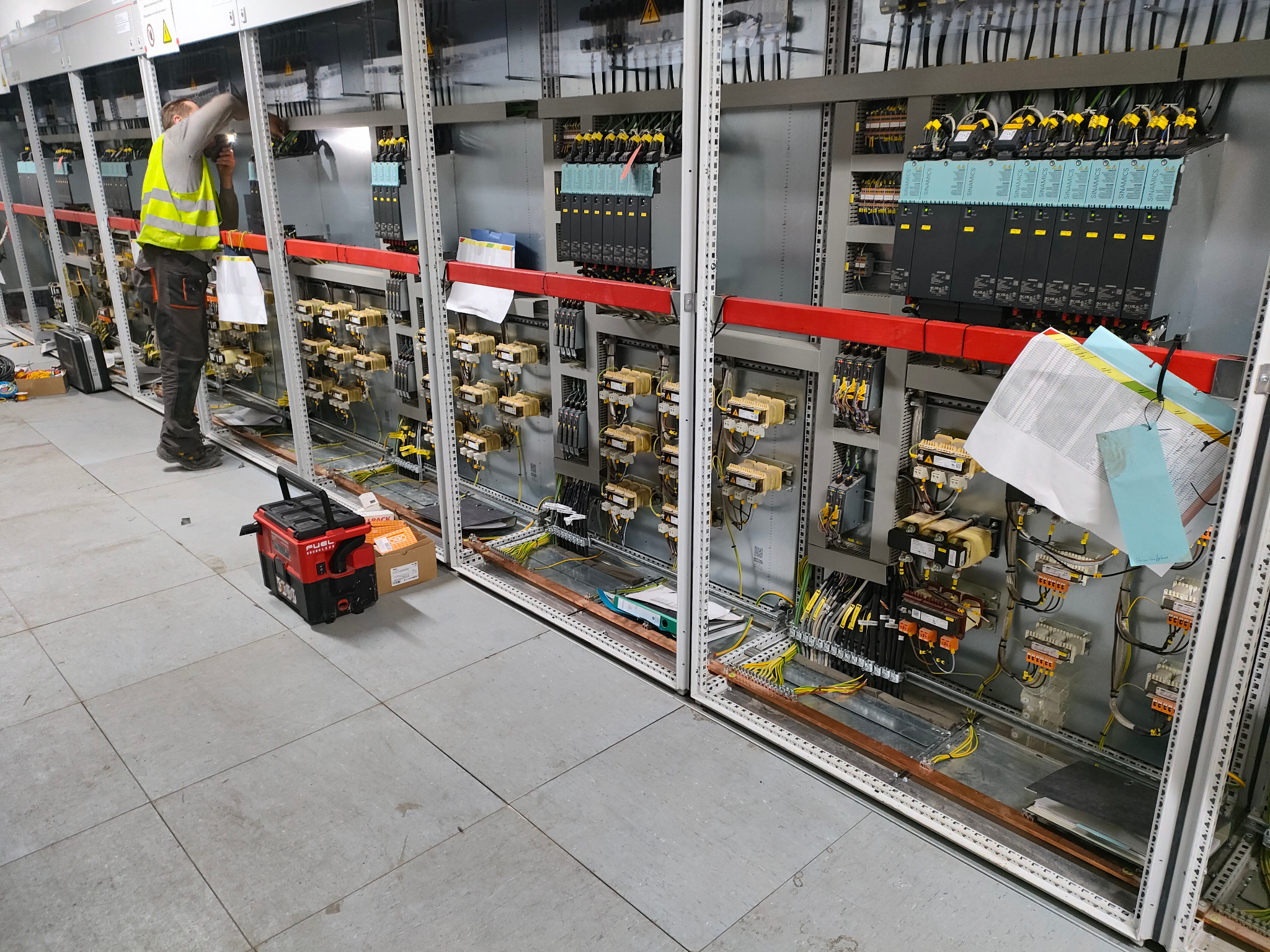 Elektryk w kamizelce odblaskowej pracuje nad rozbudowaną szafą sterowniczą z modułami Siemens Sinamics, widoczne liczne przewody, złączki, transformatory i skrzynka narzędziowa Milwaukee Fuel...