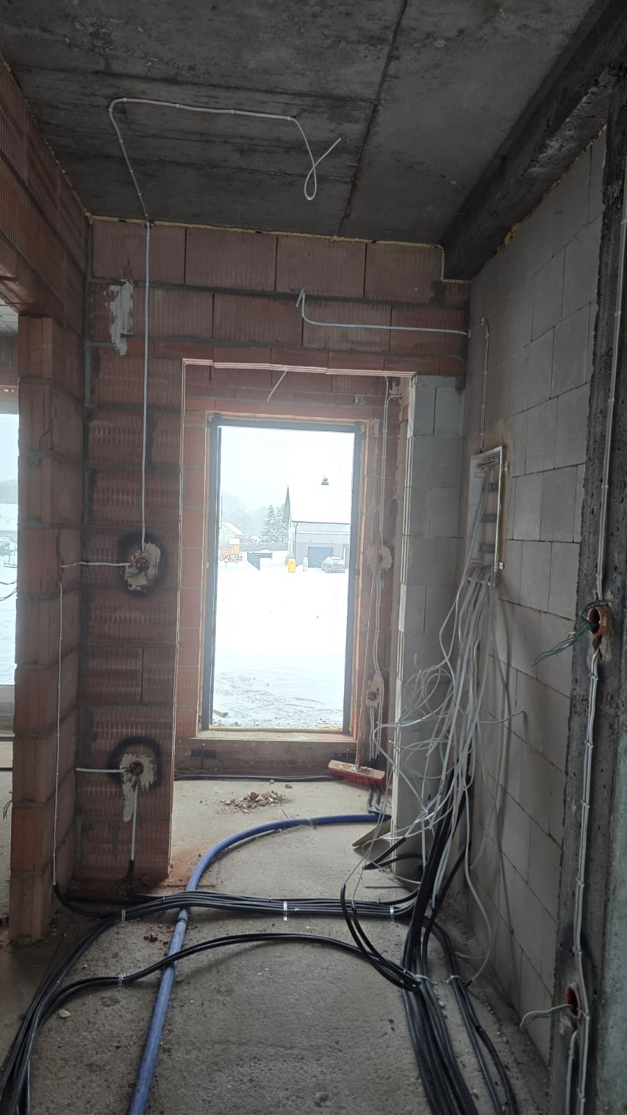 Surowy stan budynku w trakcie prac elektrycznych: kable w ścianach i na podłodze, puszki instalacyjne, widok na zewnątrz przez okno w zimowy dzień.