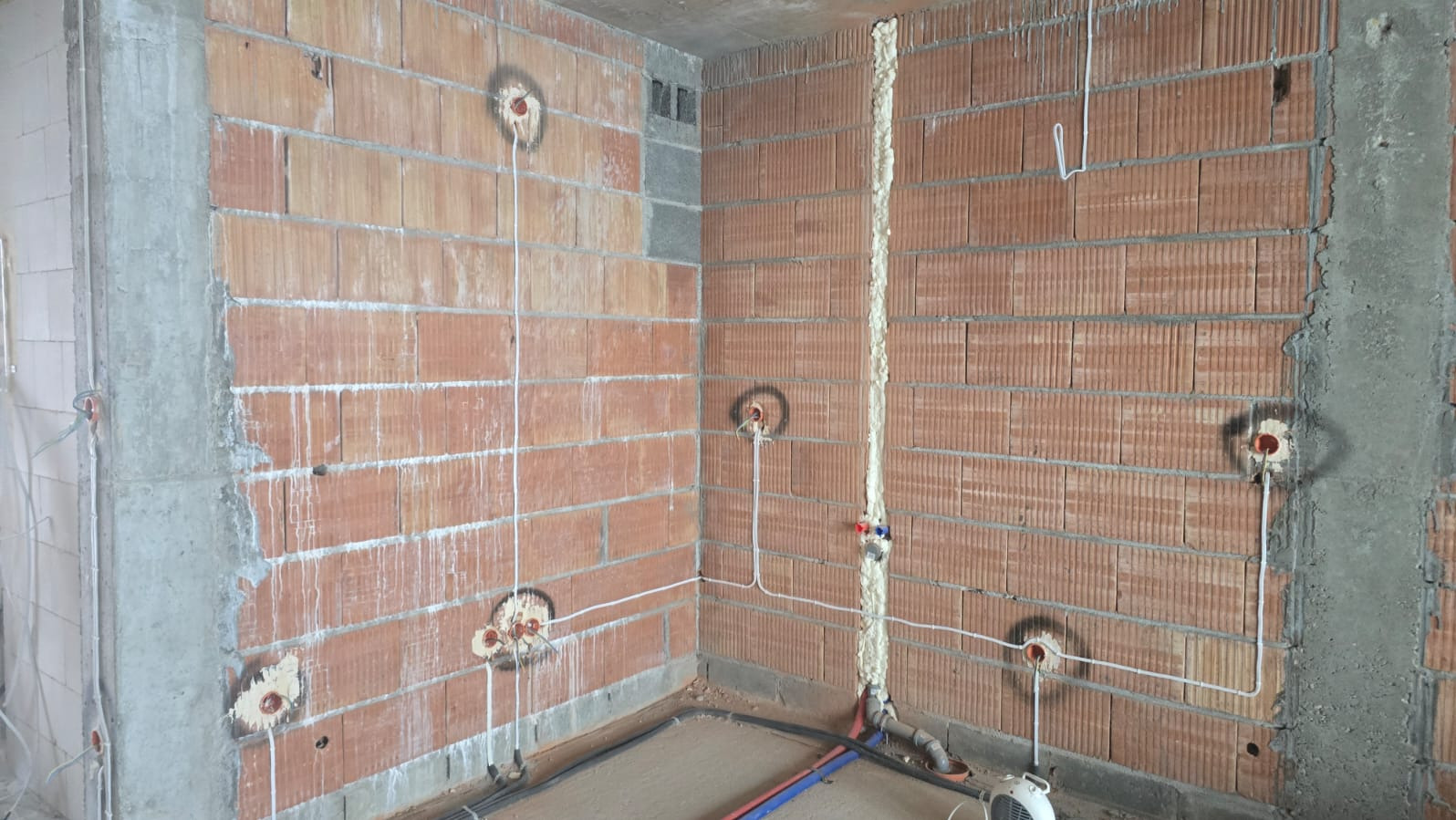 Surowy mur z widocznymi puszkami elektrycznymi i przewodami, pionowa bruzda wypełniona pianką montażową. Instalacja w trakcie realizacji, widoczne elementy hydrauliczne.