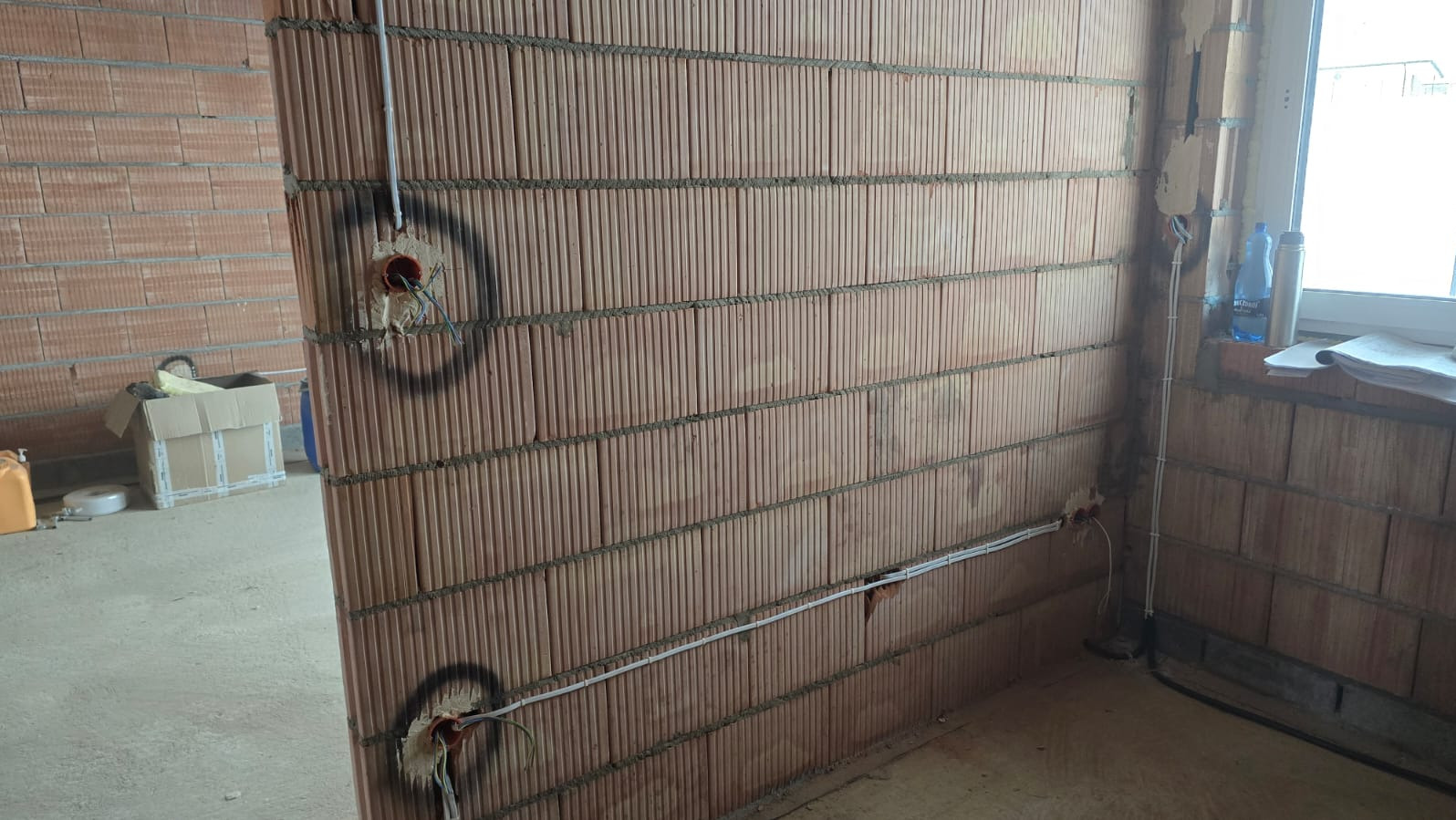 Wewnętrzna ściana z cegły z widocznymi przewodami elektrycznymi w peszlach i puszkami instalacyjnymi, przygotowana do dalszych prac wykończeniowych.