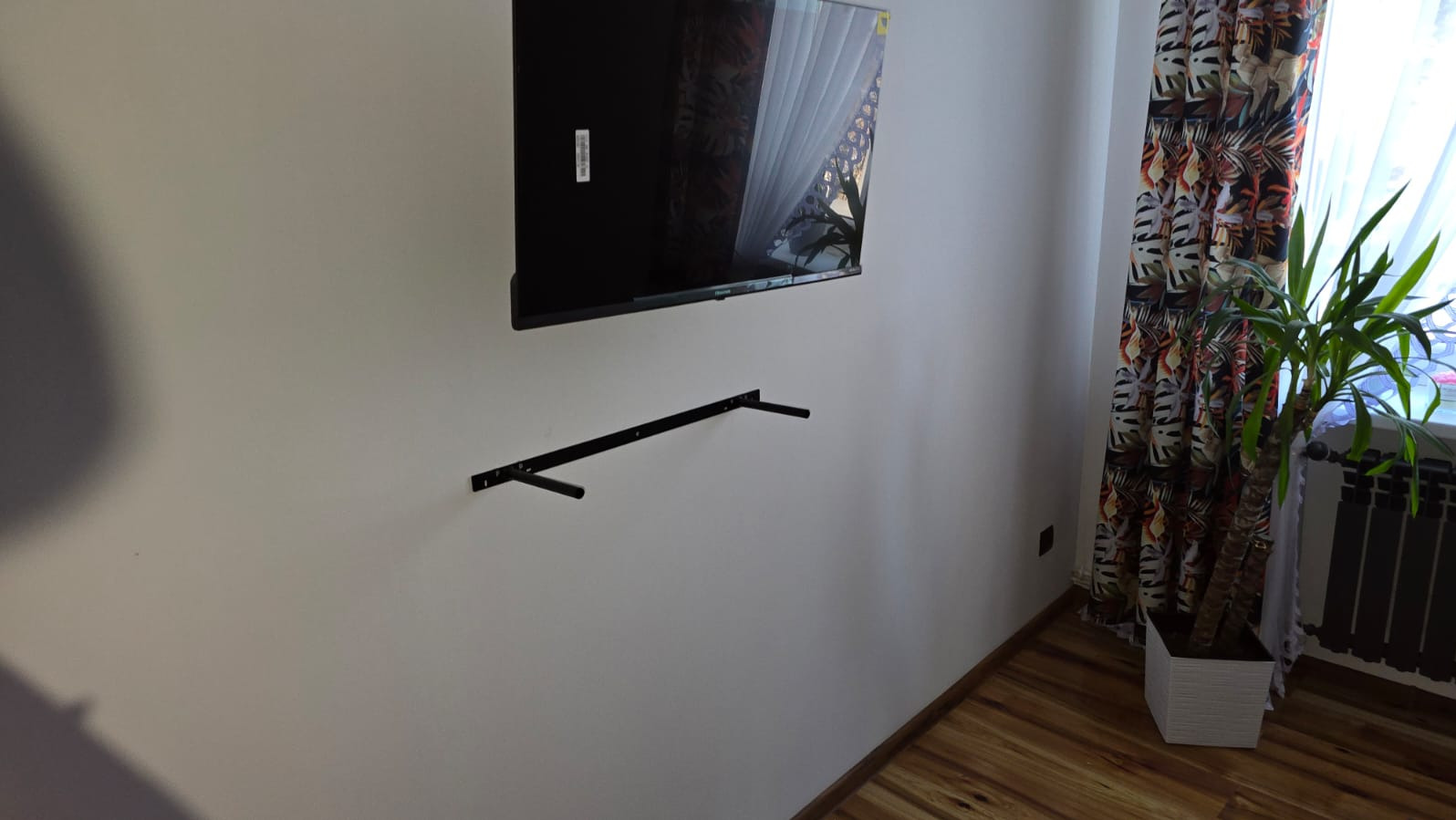 Telewizor wiszący na ścianie, zamontowany na czarnych wspornikach. W tle okno z wzorzystymi zasłonami i roślina doniczkowa. Widoczne drewniane panele podłogowe.