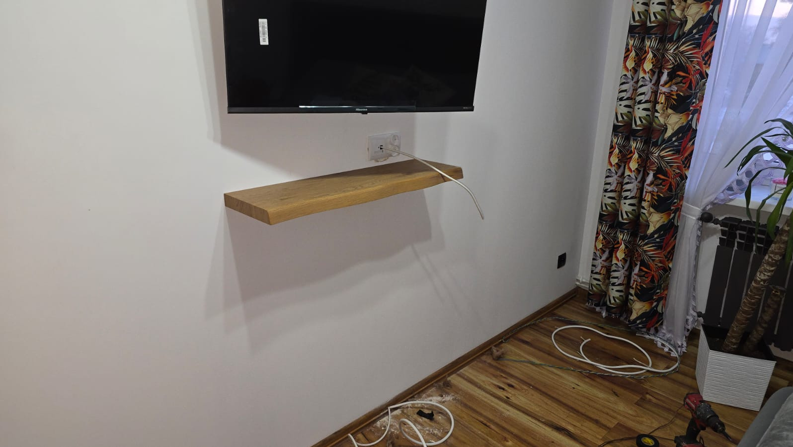 Telewizor na ścianie z drewnianą półką i gniazdkiem. Widoczne kable i narzędzia na podłodze. W tle okno z kolorowymi zasłonami i grzejnik.