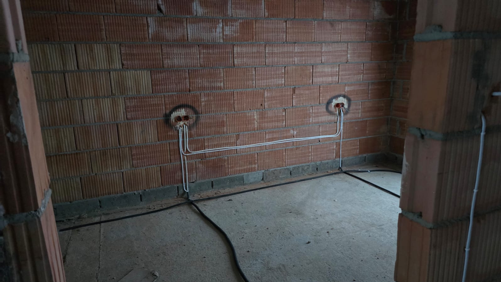 Surowa ściana z cegły z dwoma punktami elektrycznymi połączonymi białymi przewodami. Czarny kabel biegnie wzdłuż podłogi. Widok na wnętrze w trakcie prac instalacyjnych.