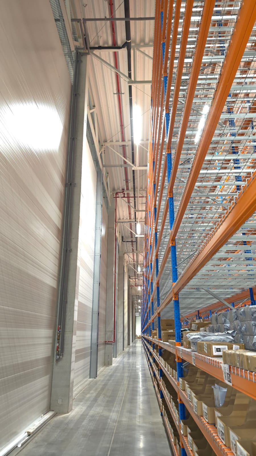 Wnętrze hali magazynowej z regałami, widoczne instalacje pod sufitem i oświetlenie. Perspektywa z dołu, podkreślająca wysokość i przestrzeń.