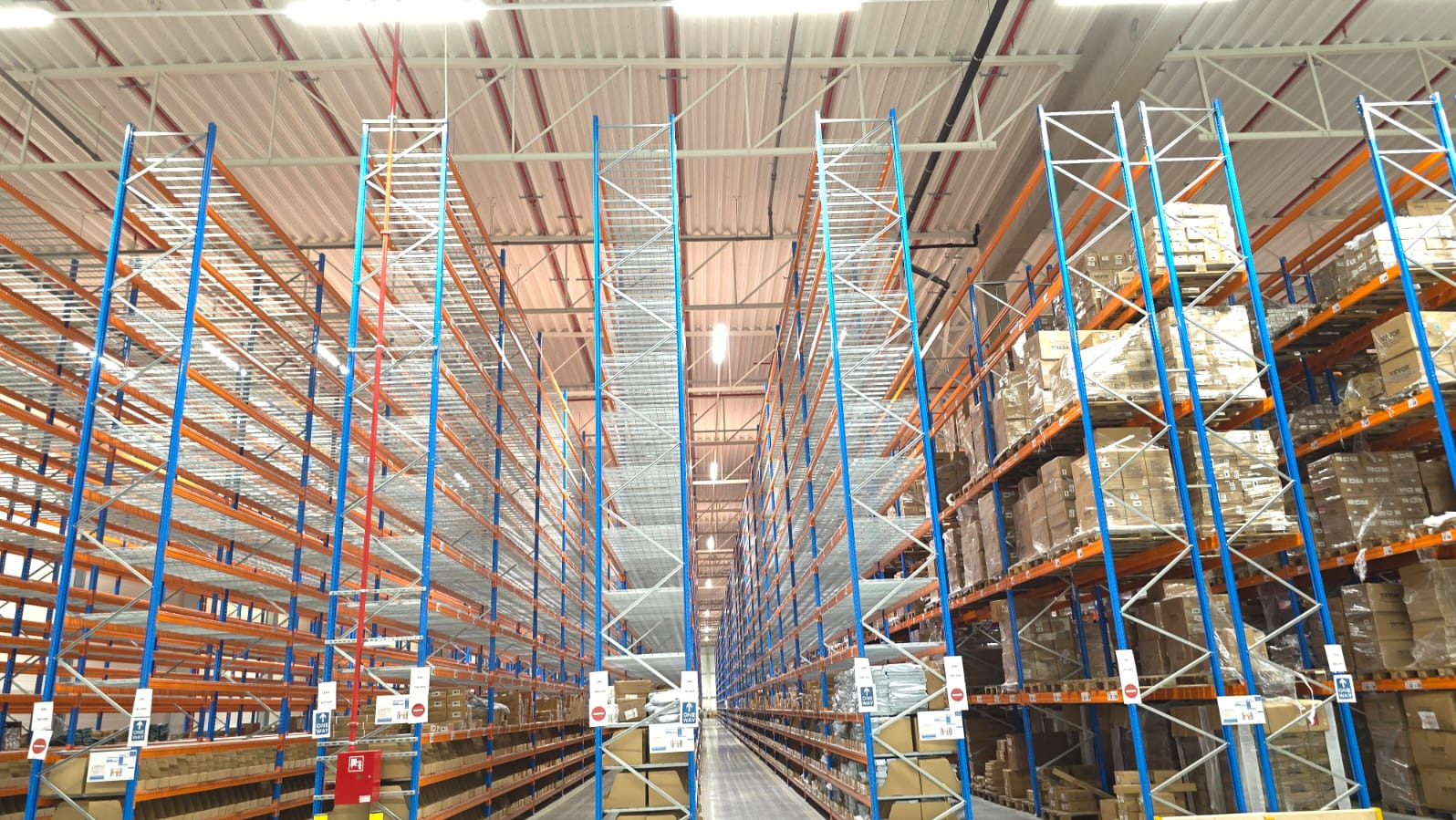 Wnętrze magazynu wysokiego składowania z regałami paletowymi, częściowo wypełnionymi kartonami. Widoczne niebieskie słupy i pomarańczowe poprzeczki regałów.