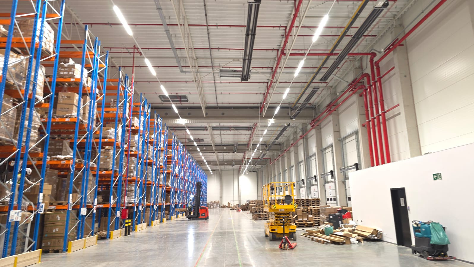 Wnętrze hali magazynowej z regałami wypełnionymi towarem, widoczne instalacje pod sufitem i maszyny do transportu towarów. Czerwone rury instalacji przeciwpożarowej.