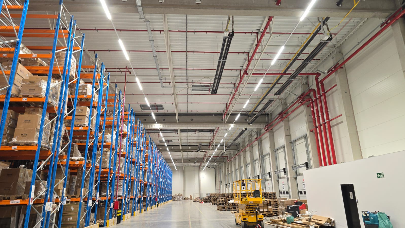 Oświetlenie LED w hali magazynowej w Jarosławiu. Widoczne regały z towarem, instalacje pod sufitem i podnośnik. Energooszczędne rozwiązanie dla przemysłu.